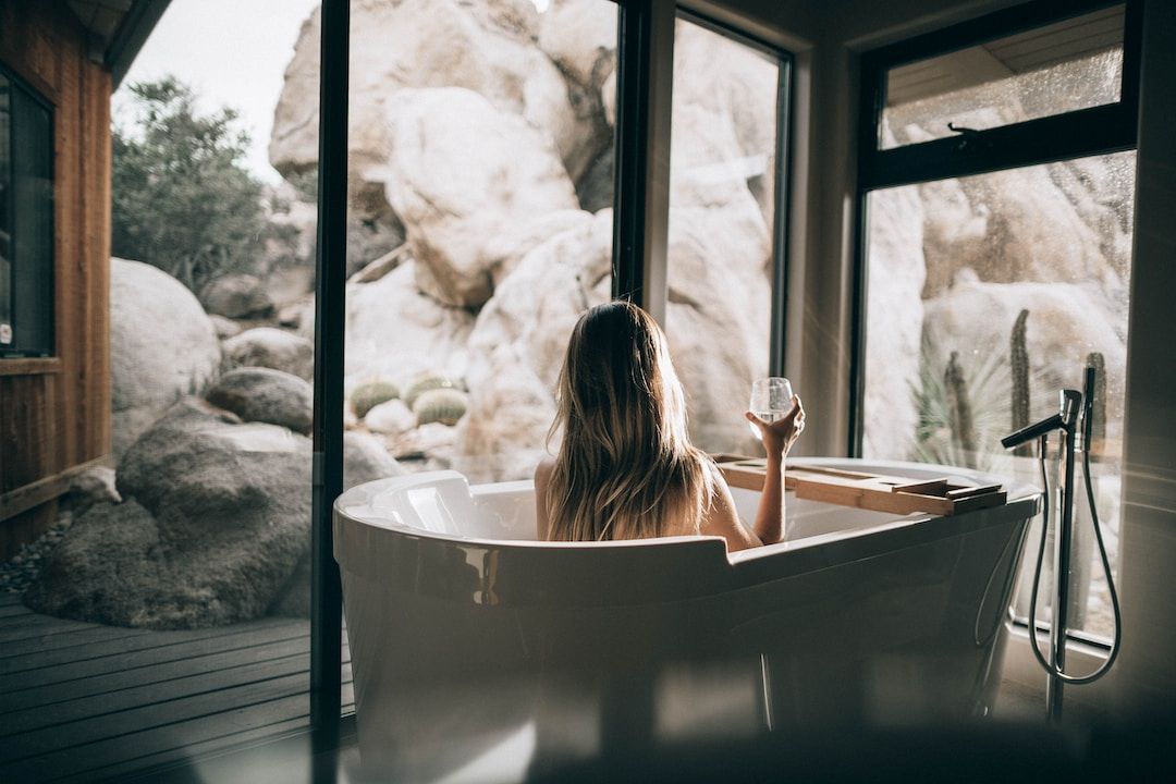 A woman relaxing in a freestanding bathtub by a large window overlooking rocks and trees