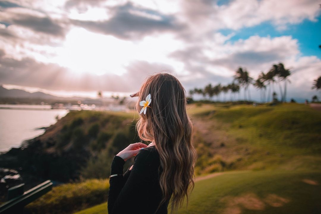 A woman with long hair standing on a grassy hill looking out at the sky