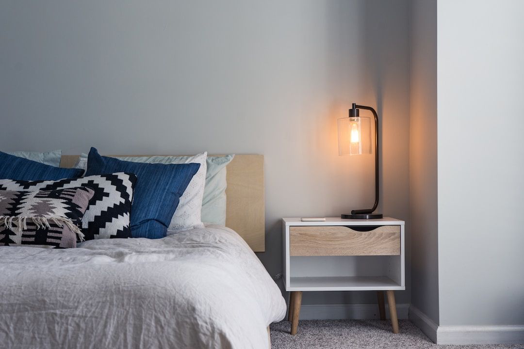 A neatly made bed with blue pillows beside a nightstand and a warm lamp