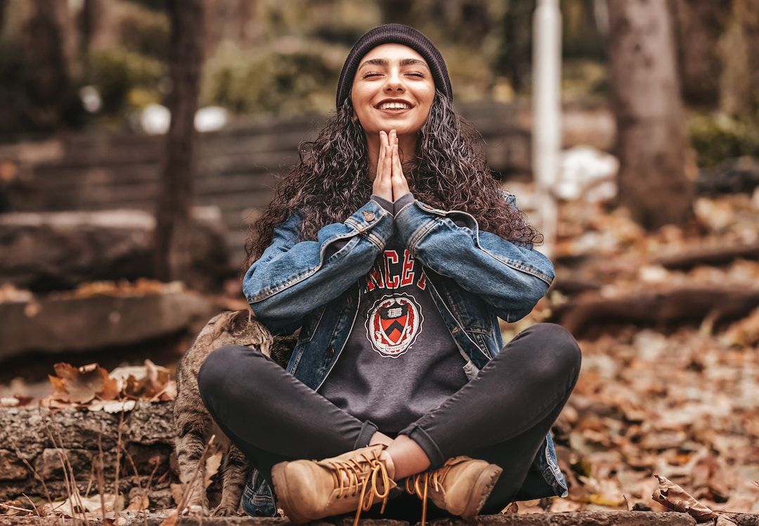 A smiling woman sitting cross-legged in the woods with her palms together