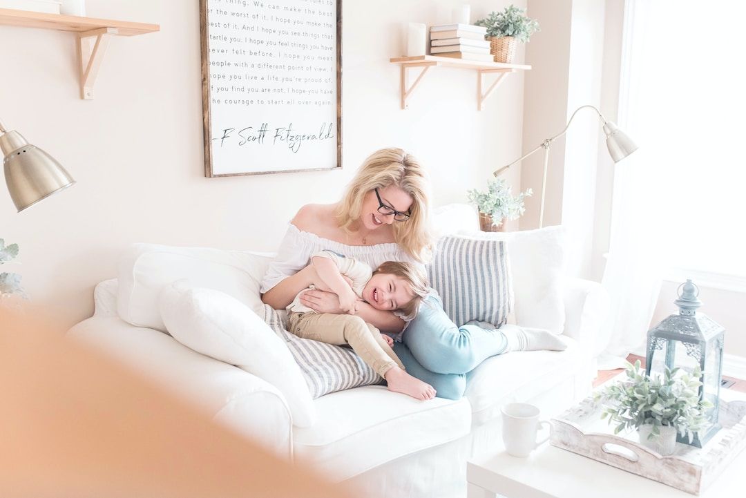 A mother lying on a white sofa reading with her young child in a bright room
