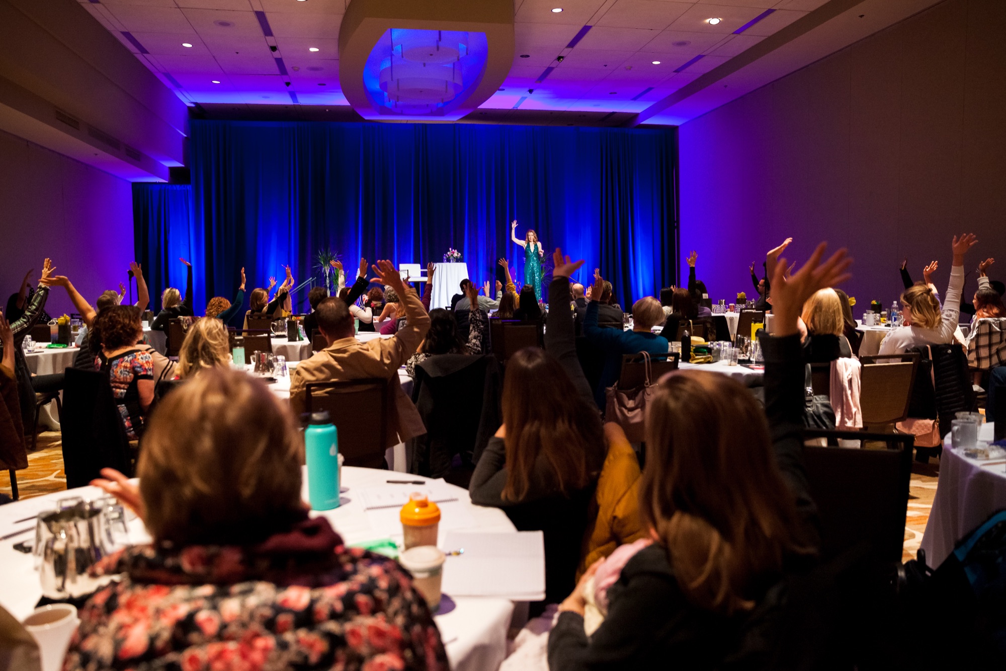 A ballroom of women with their hands raised as Tamara Rose leads from the stage