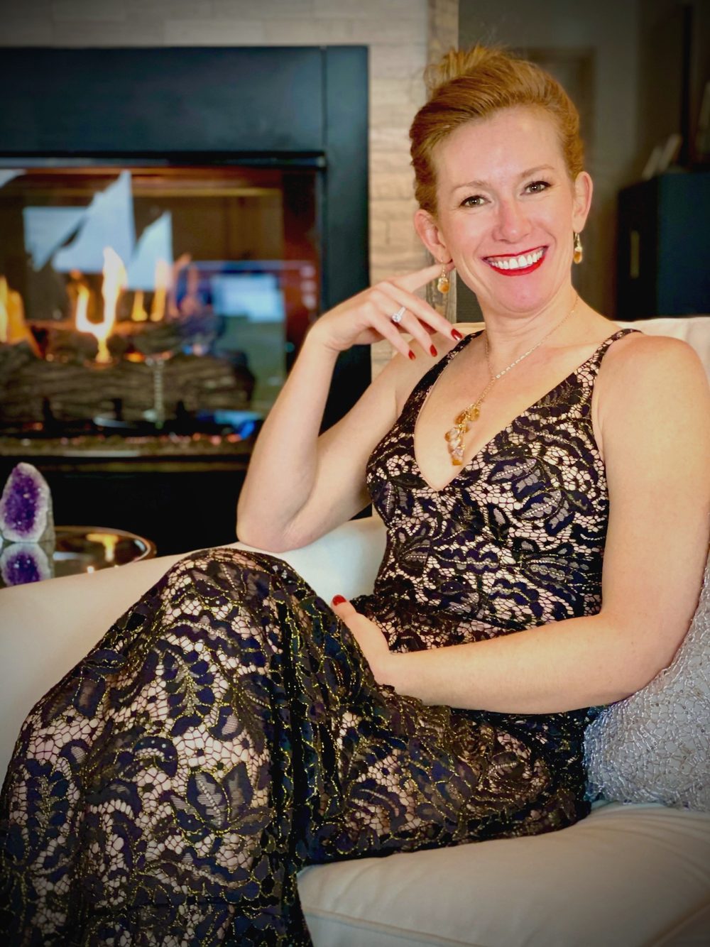 Tamara Rose smiling in a black lace dress, seated by a fireplace
